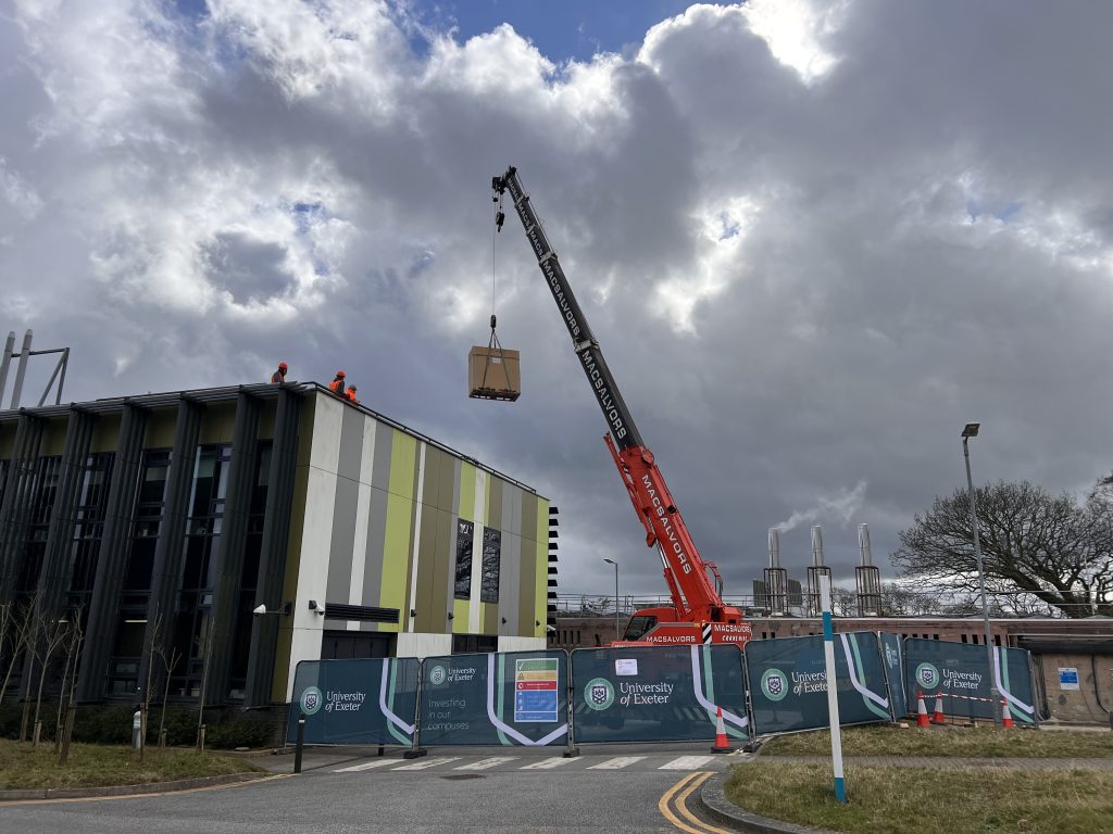 A photograph of a crane lifting a large boxed air source heat pump being crane lifted into the air in front of the Stella Turk building at Penryn campus.