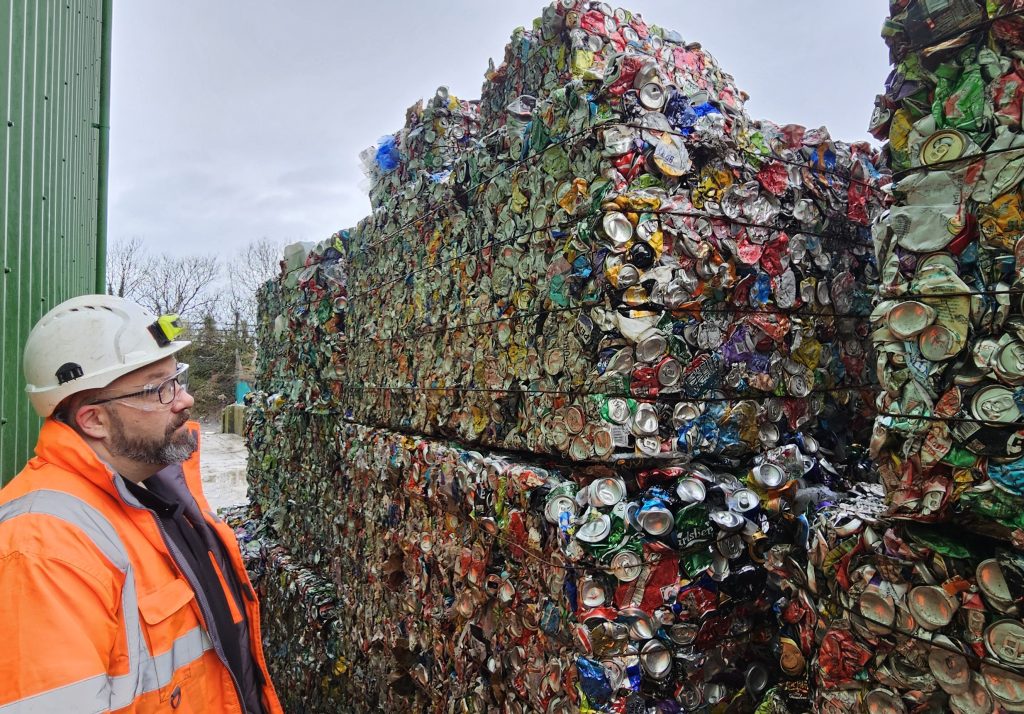 Man in a high-vis jacket and white hard hat looking at blocks of compacted tins and cans towered up.