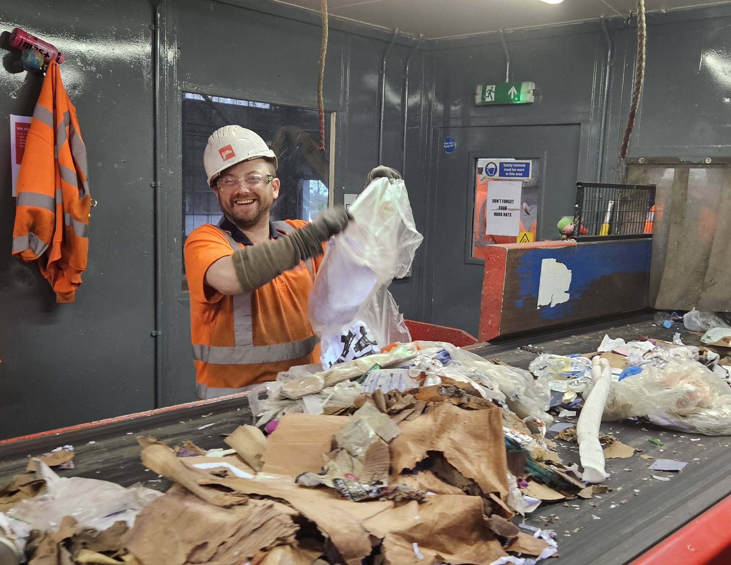A male in a orange high-vis jacket, white hard hat, protective glasses and gloves, sorting waste and recycling on a conveyor belt.