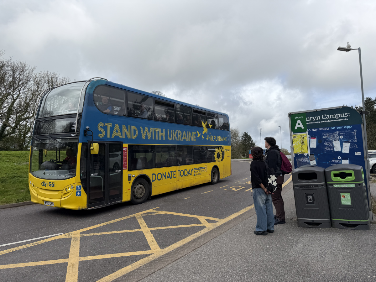 image of a Go Cornwall bus driving through Penryn campus bus stop with students waiting in the bus shelters.