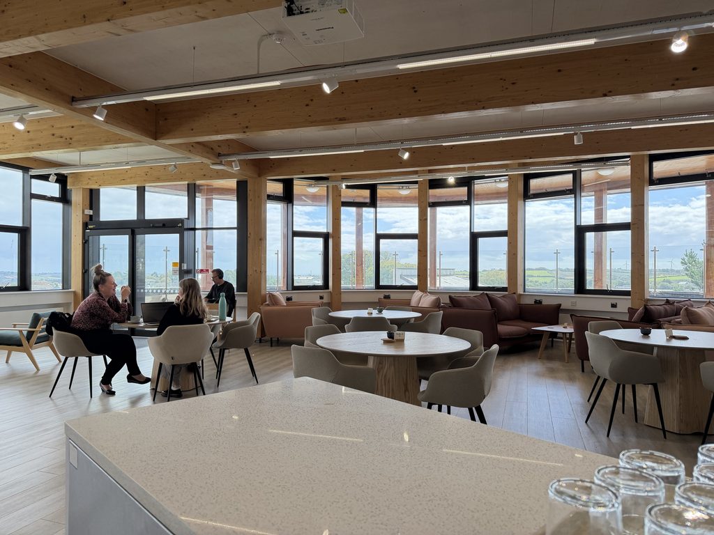 Another photo showing the newly refurbished Sustainability Cafe, looking out across a white granite counter at round wooden tables with upholstered bucket seats in neutral tones. There are some students and staff sat at tables, on laptops with cups of coffee and conversing.