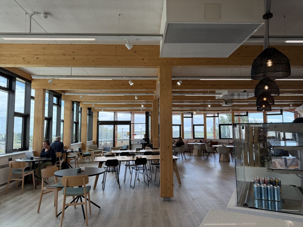 A photo of the refreshed Sustainability Cafe refurbishments, with new flooring, and new sustainable furniture made of light wood and salmon coloured sofas. The space is bright and airy, with panoramic views looking out across Penryn.