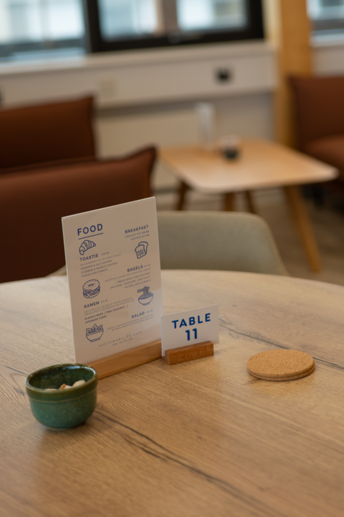 A decorative photo of the Sustainability cafe table menu on a wooden table with a coaster and pot of sugar cubes next to it.