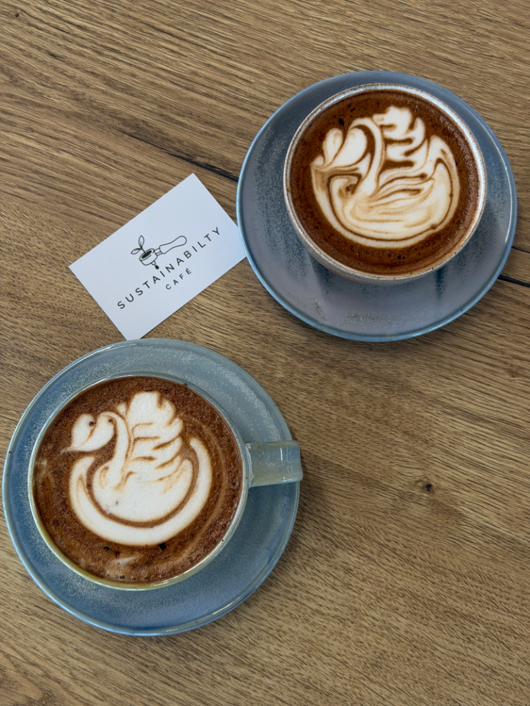 Image of the Sustainability Cafe's new branding on a loyalty card between two barista made coffees with coffee art showing swans and patterns in the foam.